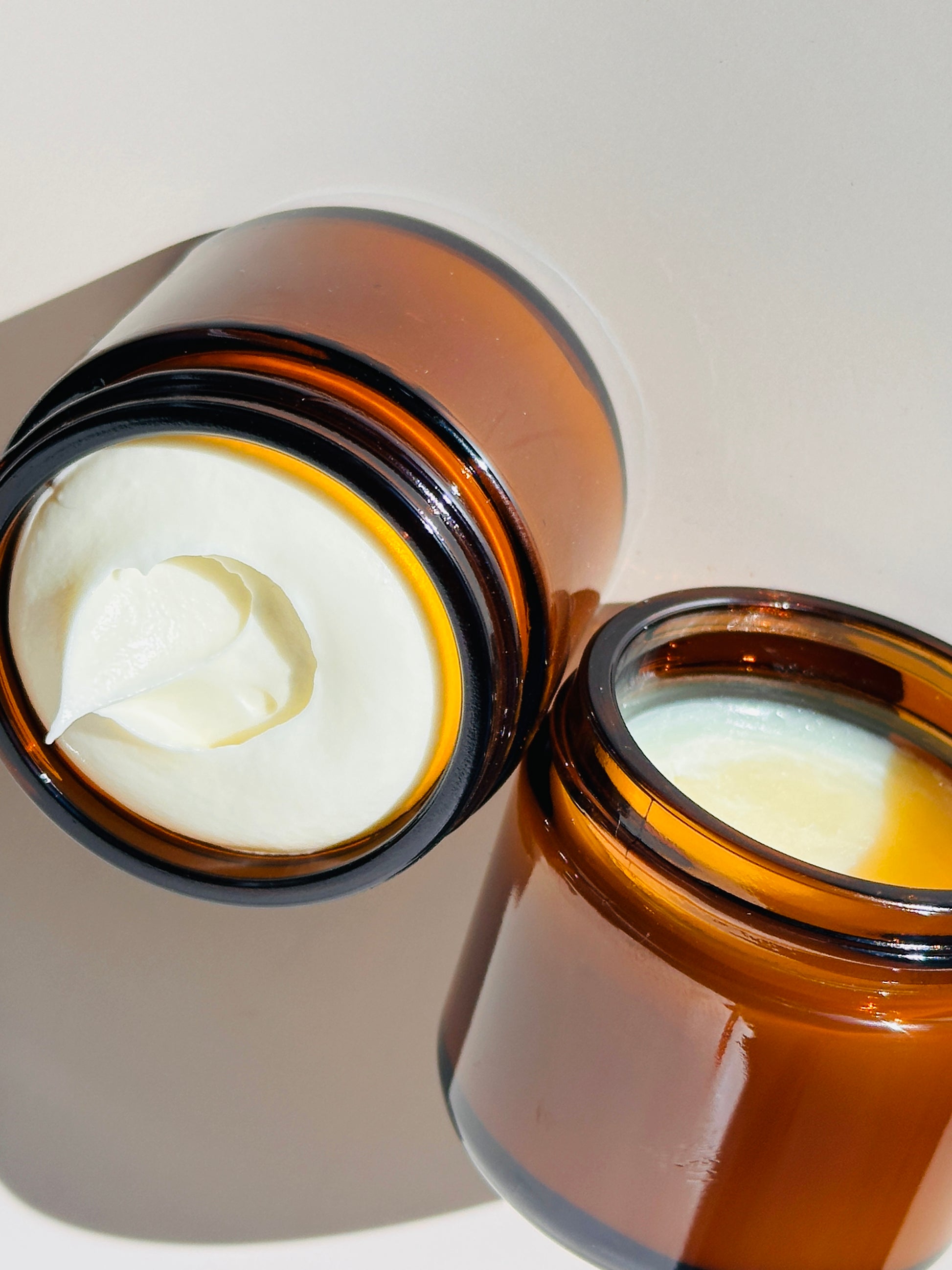 Two open brown glass jars filled with white cream on a neutral background.