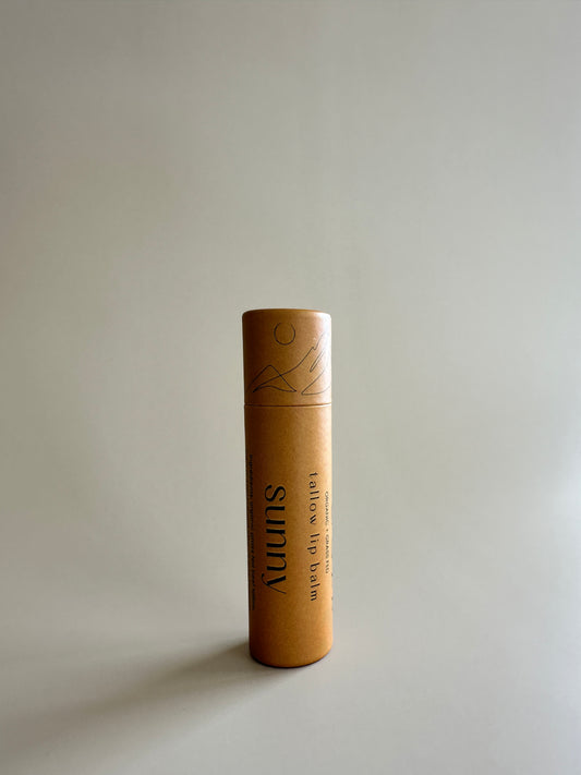 A cardboard tube of lip balm with a natural beige color, placed against a neutral background.