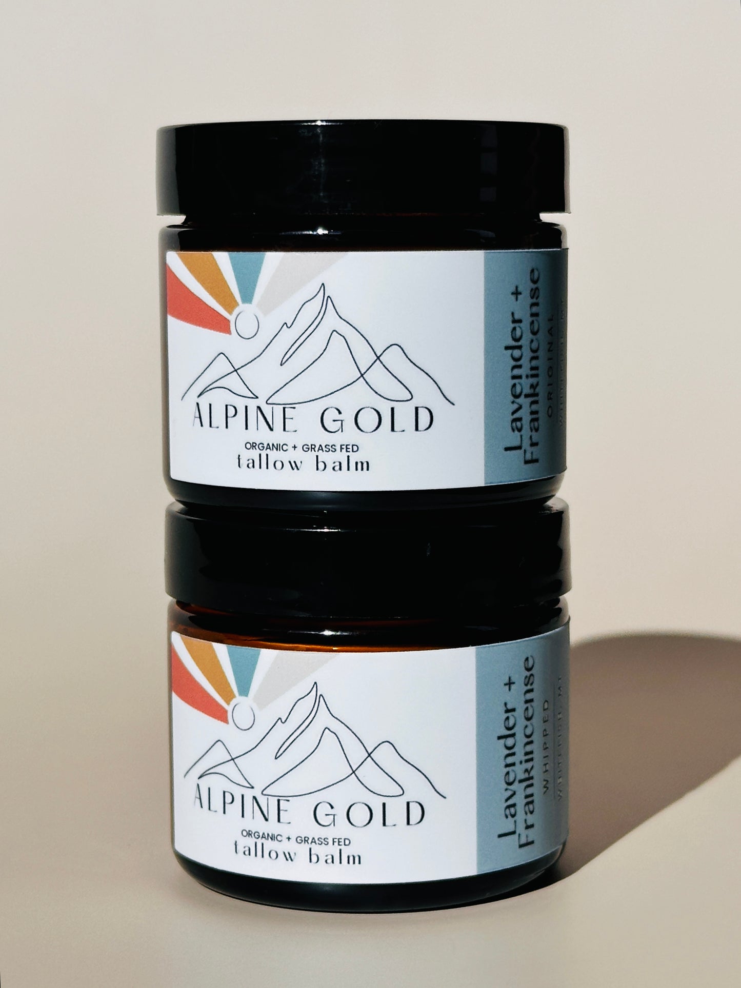 Two jars of Alpine Gold tallow balm with mountain logo and colorful label on neutral background