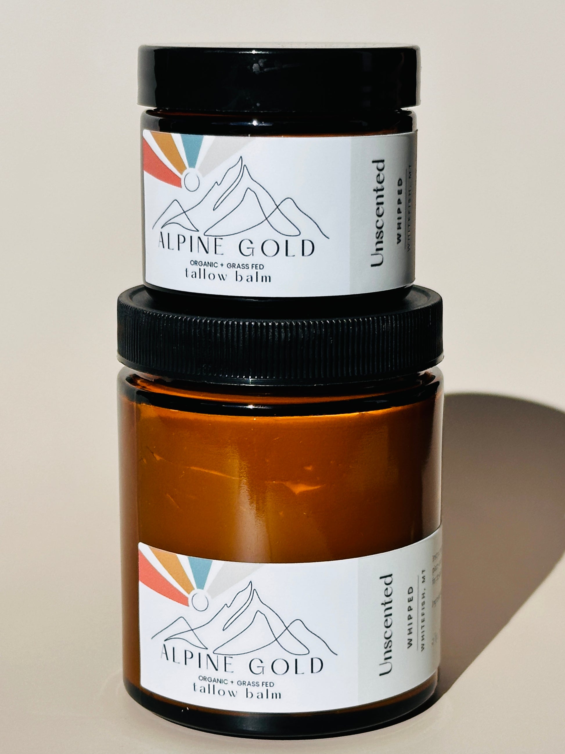 Two jars of organic Alpine Gold tallow balm on a neutral background. Unscented tallow balm in 2oz and 6oz.