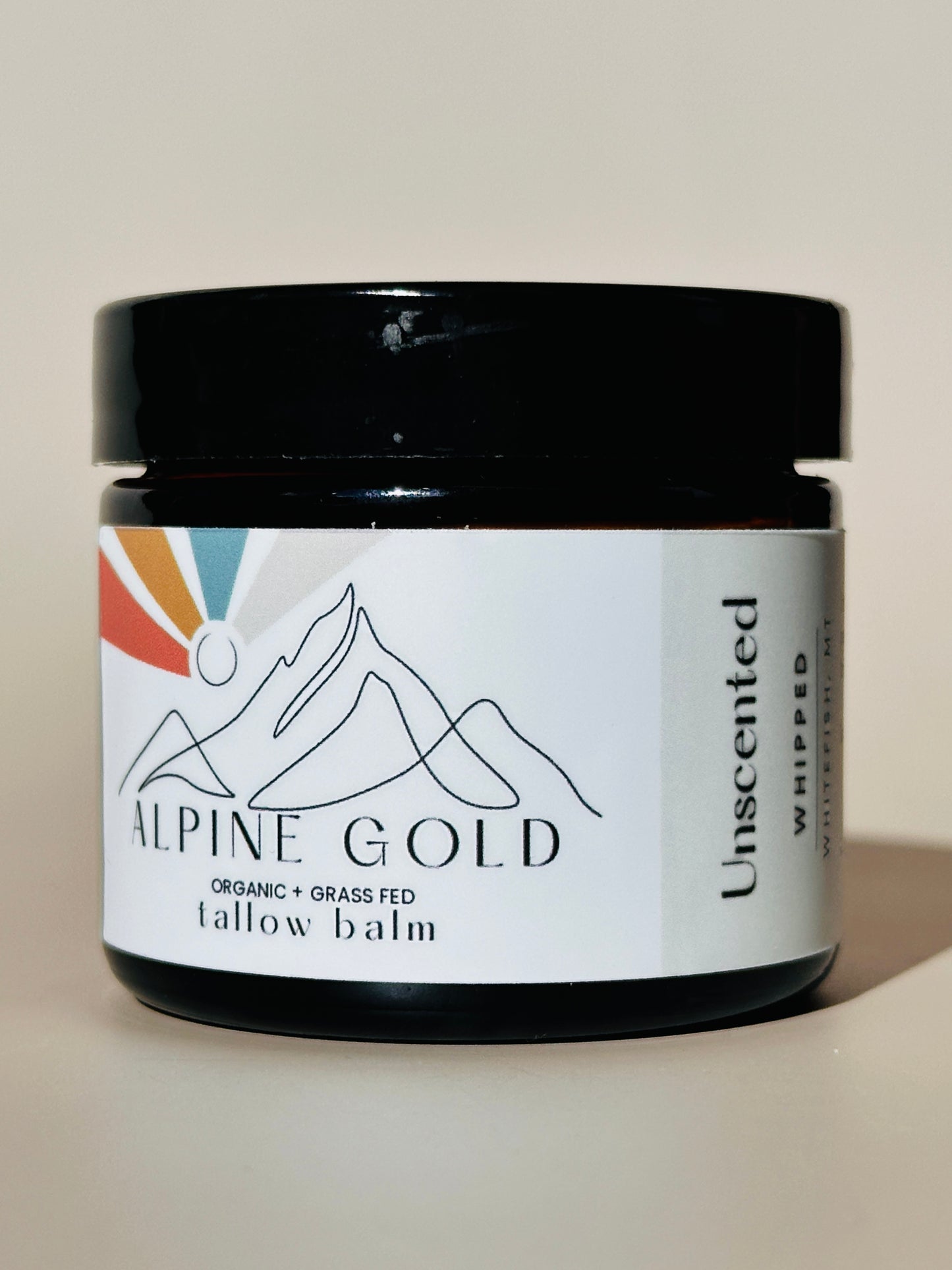 A jar of ALPINE GOLD Tallow Balm with a label showing the brand name, product name, and an illustration of mountains. The jar is black, and the label has a white background with black and multicolor elements.